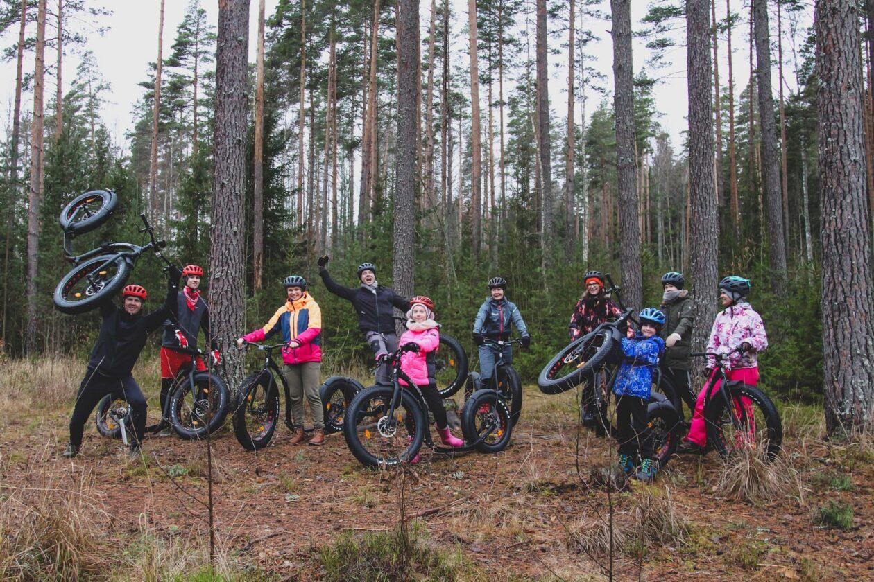 FAT-tõukerattamatkad Lõuna-Eestis. Grupp matkajaid on kogunenud fotole, FAT tõukerattad rõõmu märgiks osaliselt üles tõstetuna. Koolinoored ja täiskasvanud kõik koos lustlikult mändide all. On sügistalv.